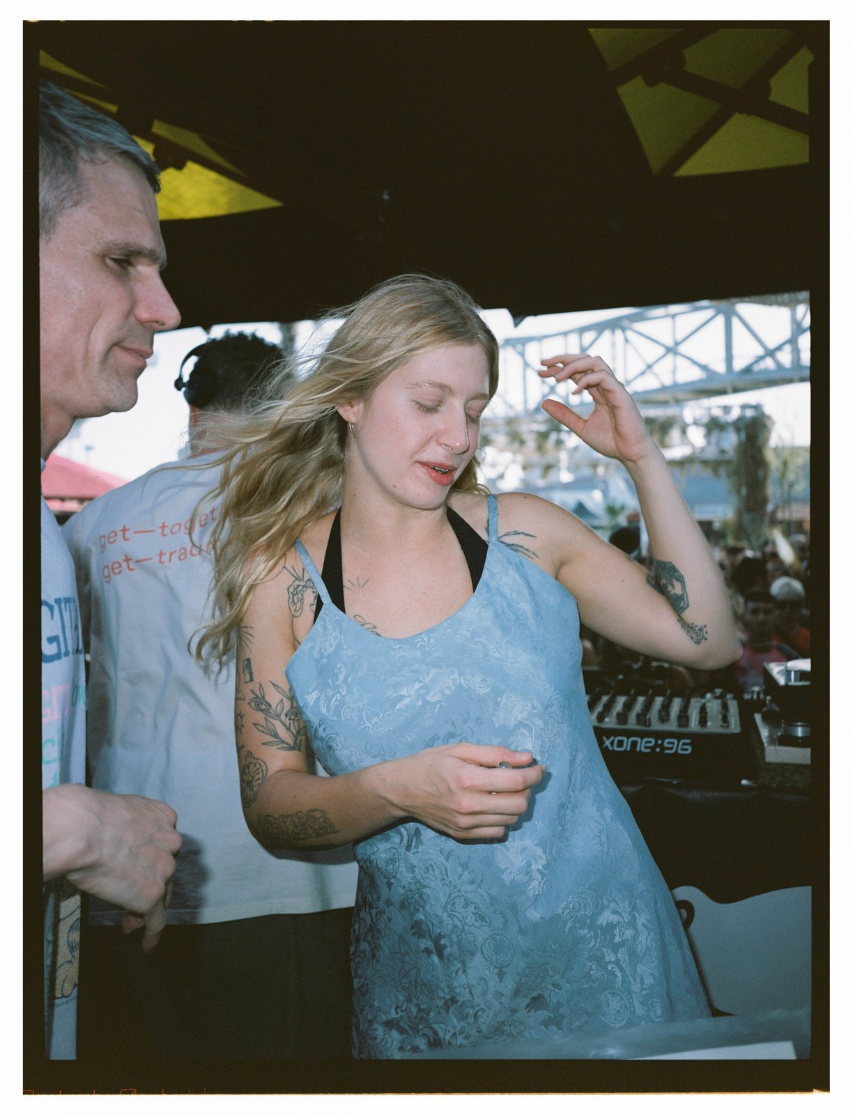 A blonde woman in a blue lace dress dances with eyes closed beside a man in a printed tee, mixer and crowd behind.