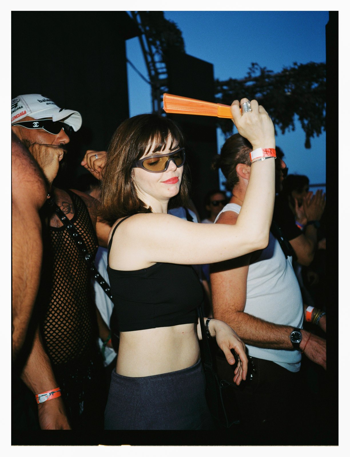 Dusk flash of a woman in orange-lens sunglasses dancing with an orange fan raised overhead in a crowd.
