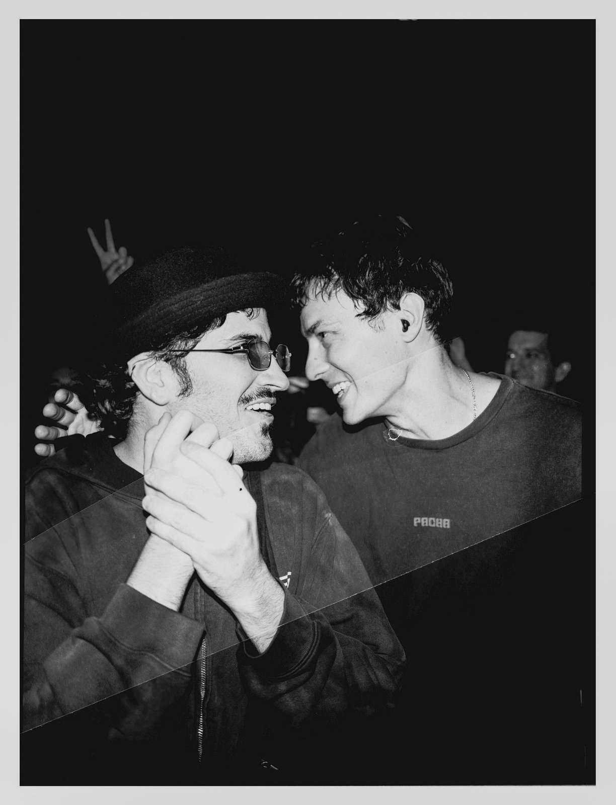Black-and-white flash shot of two grinning men leaning forehead-to-forehead, one in a bowler hat and sunglasses, crossed by a diagonal light leak.