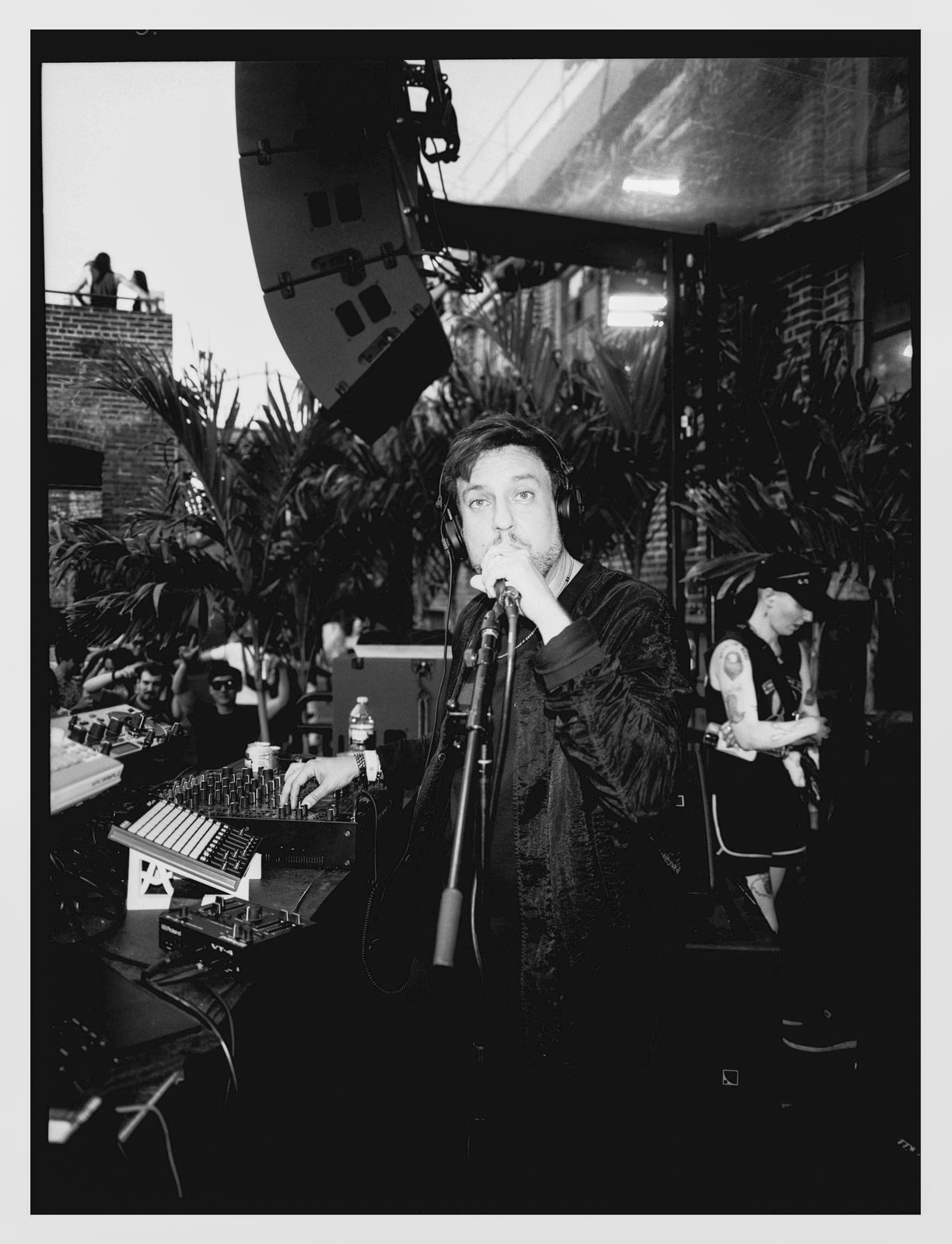 Black-and-white shot of a man in headphones singing into a mic over a synth, speakers behind on a rooftop.