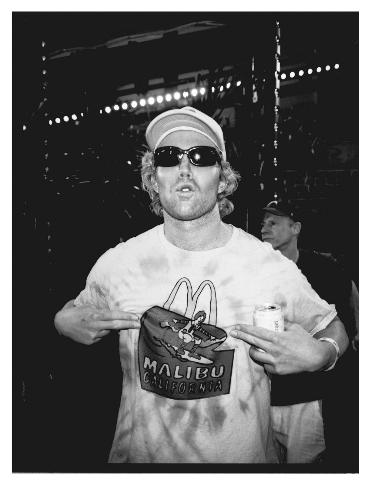 Grainy black-and-white flash portrait of a man in a cap and sunglasses stretching a tie-dye T-shirt while holding a can.