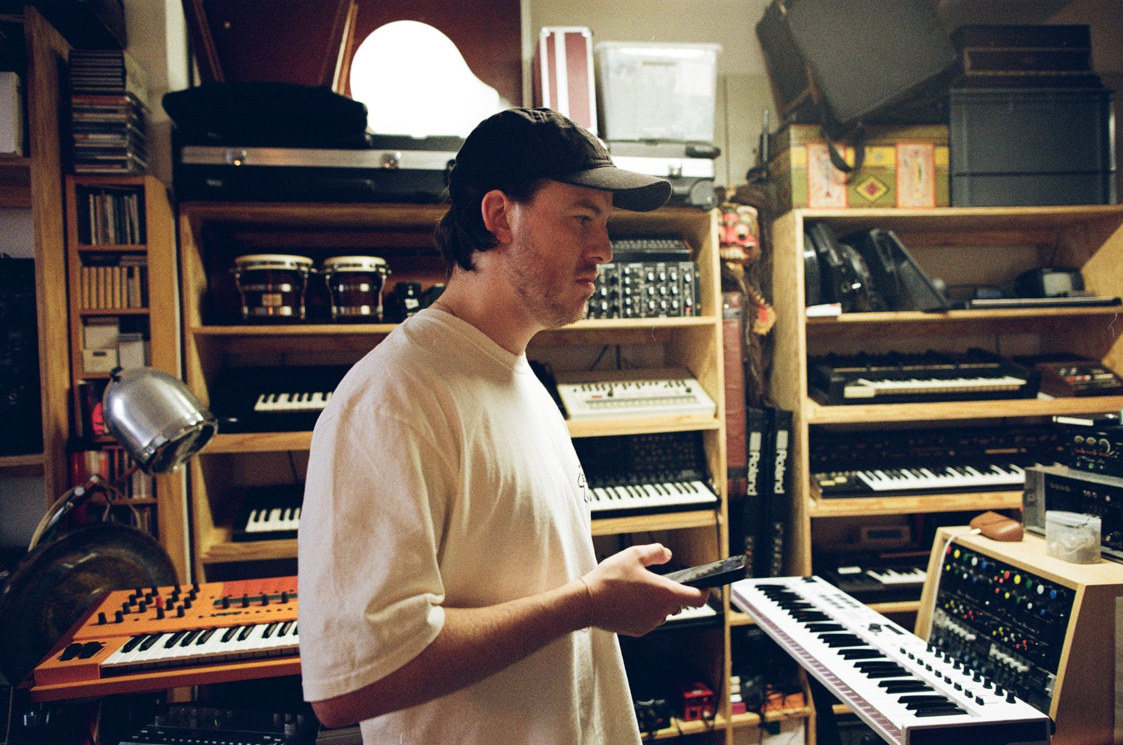 A man in a cap and white tee stands among shelves of synthesizers and keyboards in a studio.