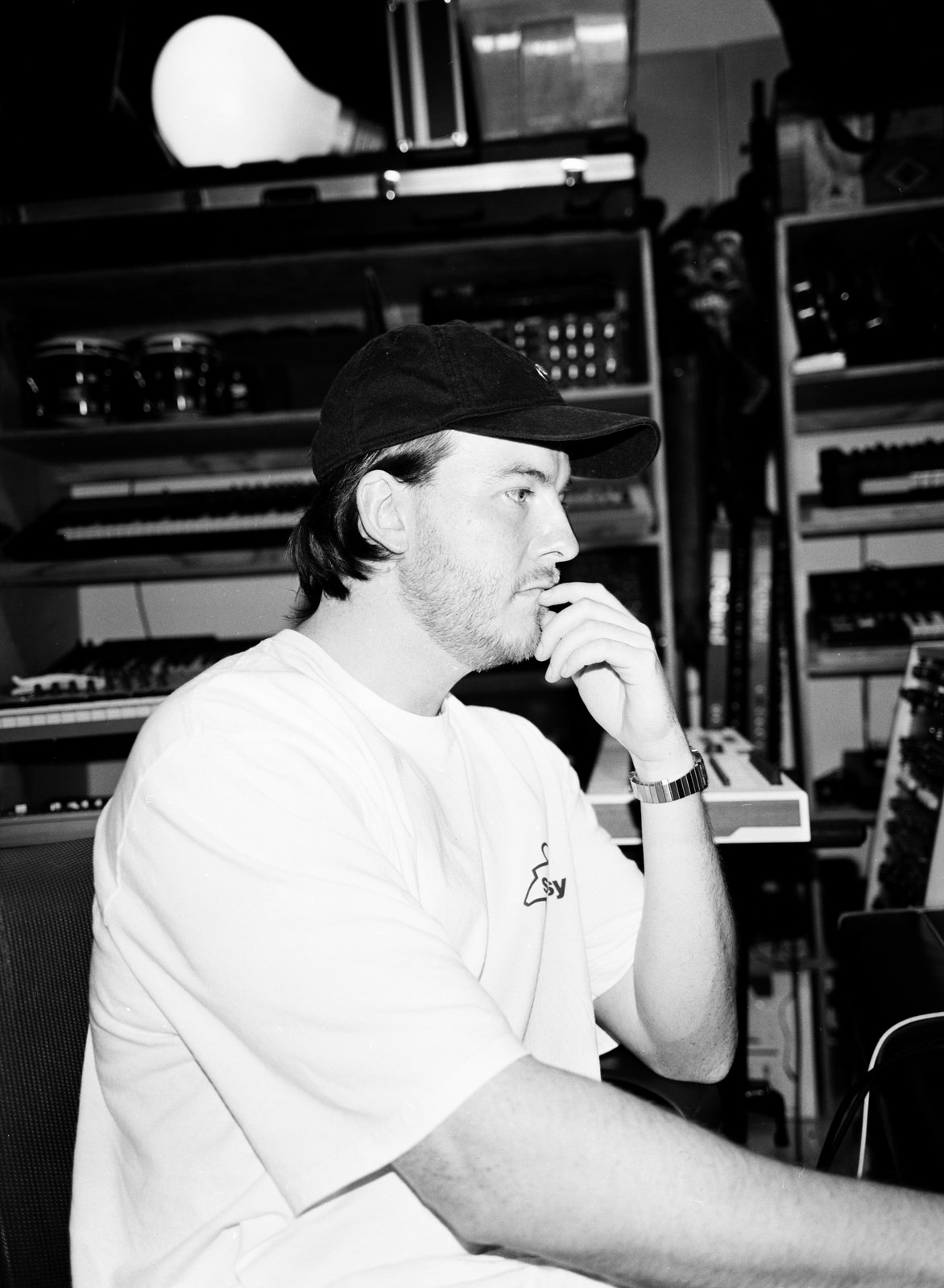 Black-and-white profile of a pensive man in a cap resting his chin, surrounded by studio gear.