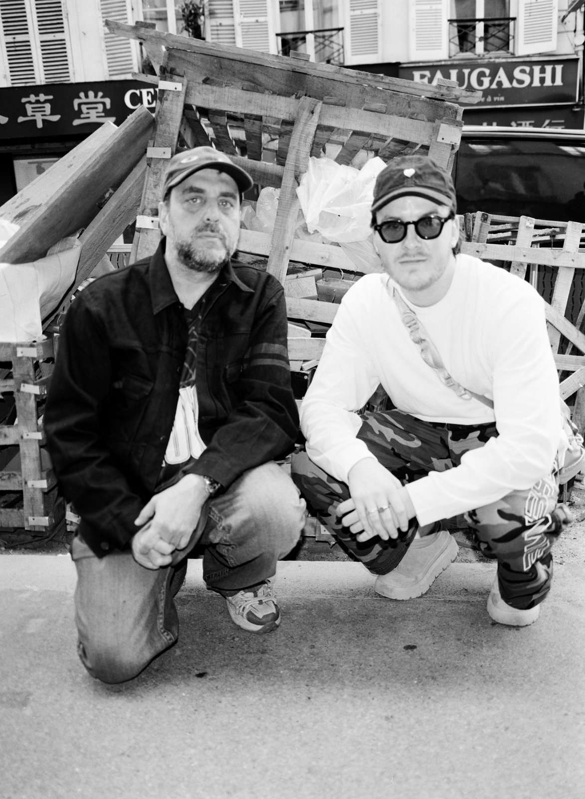 Black-and-white shot of two men in caps crouching before wooden pallets on a city street.