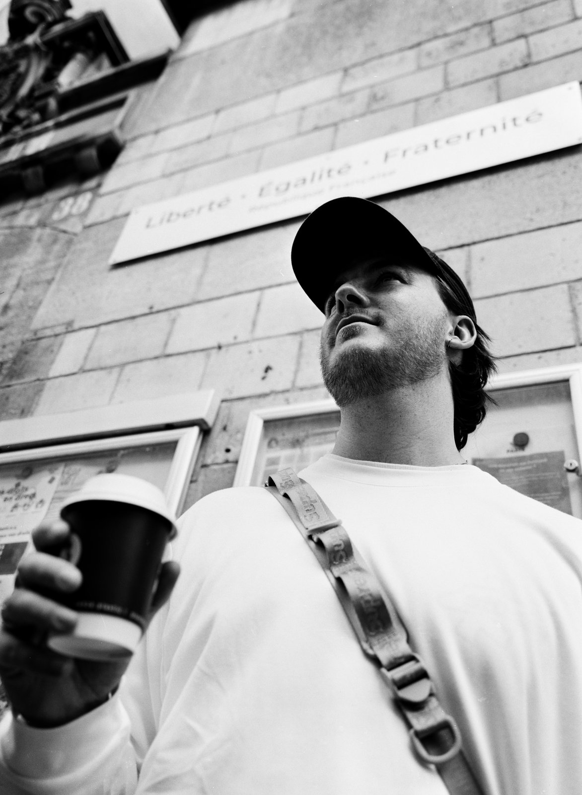Black-and-white low-angle of a man in a cap holding coffee below a street sign.