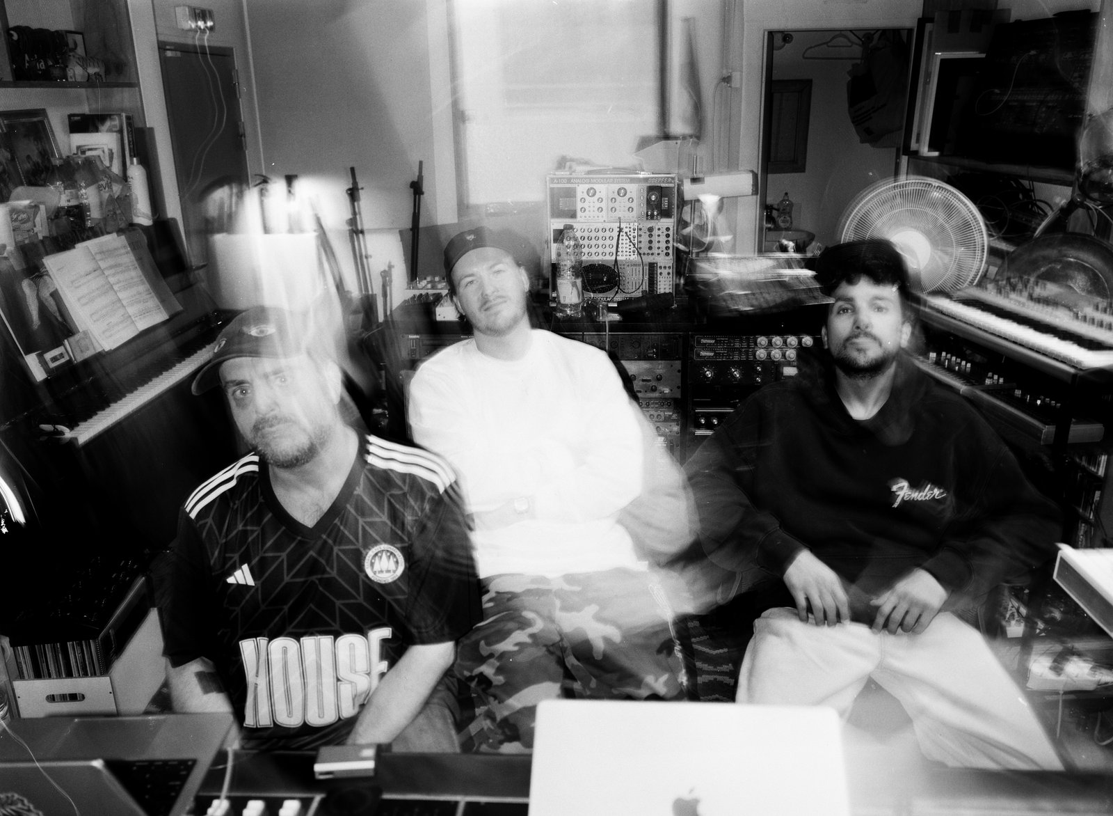 Black-and-white blurred long-exposure of three men seated in a studio full of keyboards and gear.