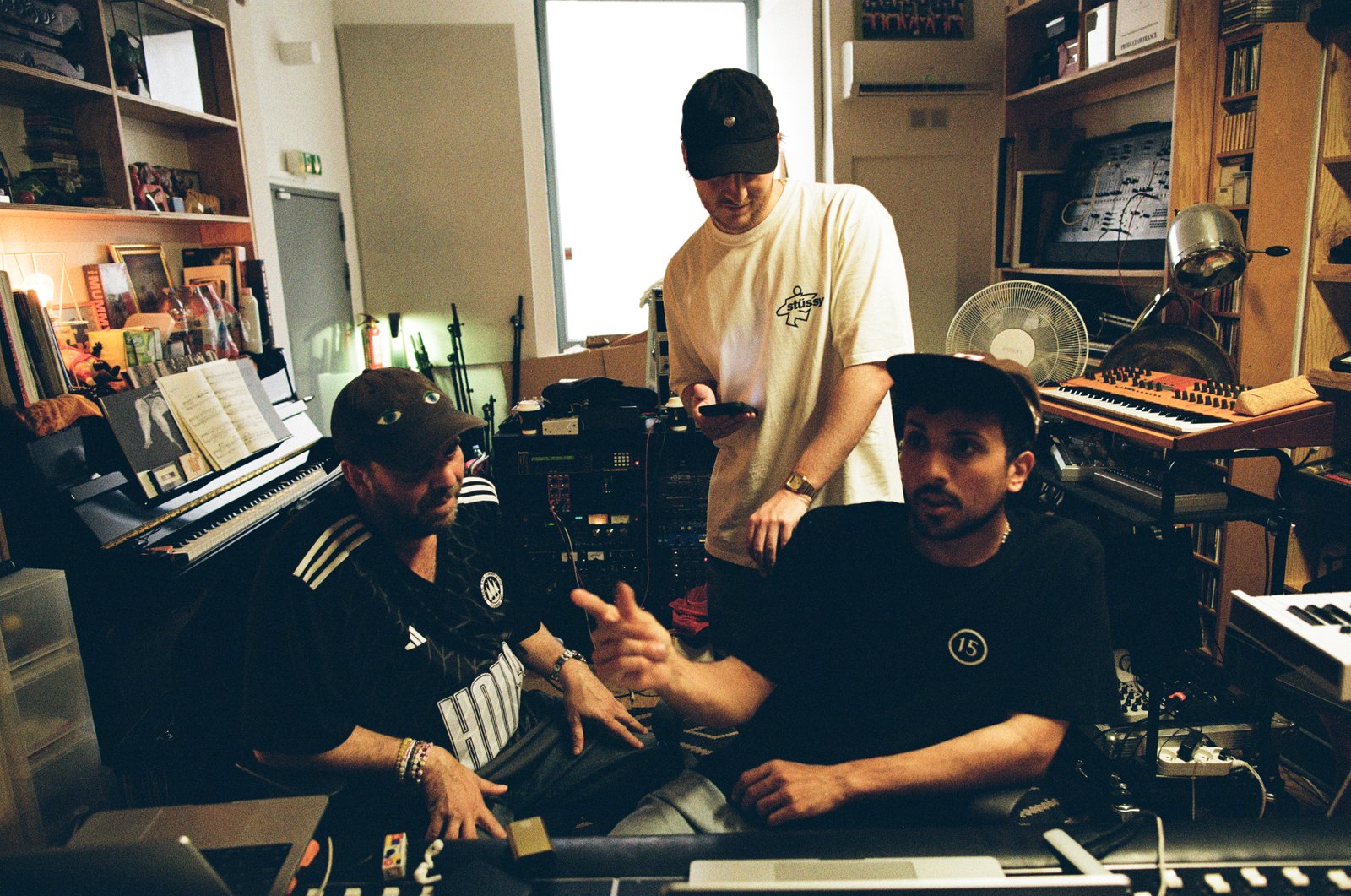 Three men gather amid keyboards and equipment in a cluttered recording studio, warm available light.