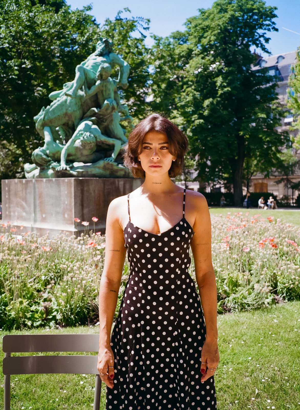 A woman in a black polka-dot slip dress stands before a green bronze statue in a sunlit park.