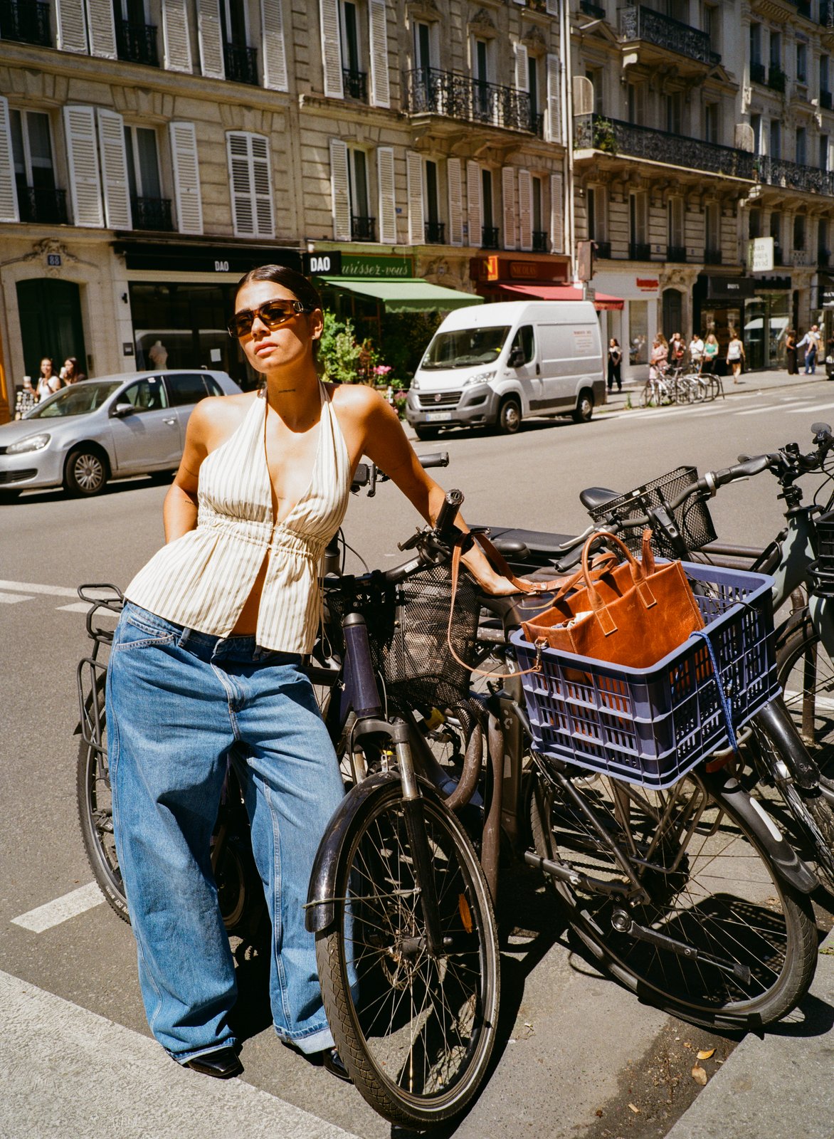 A woman in a striped top and baggy jeans leans against parked bicycles on a sunny city street.