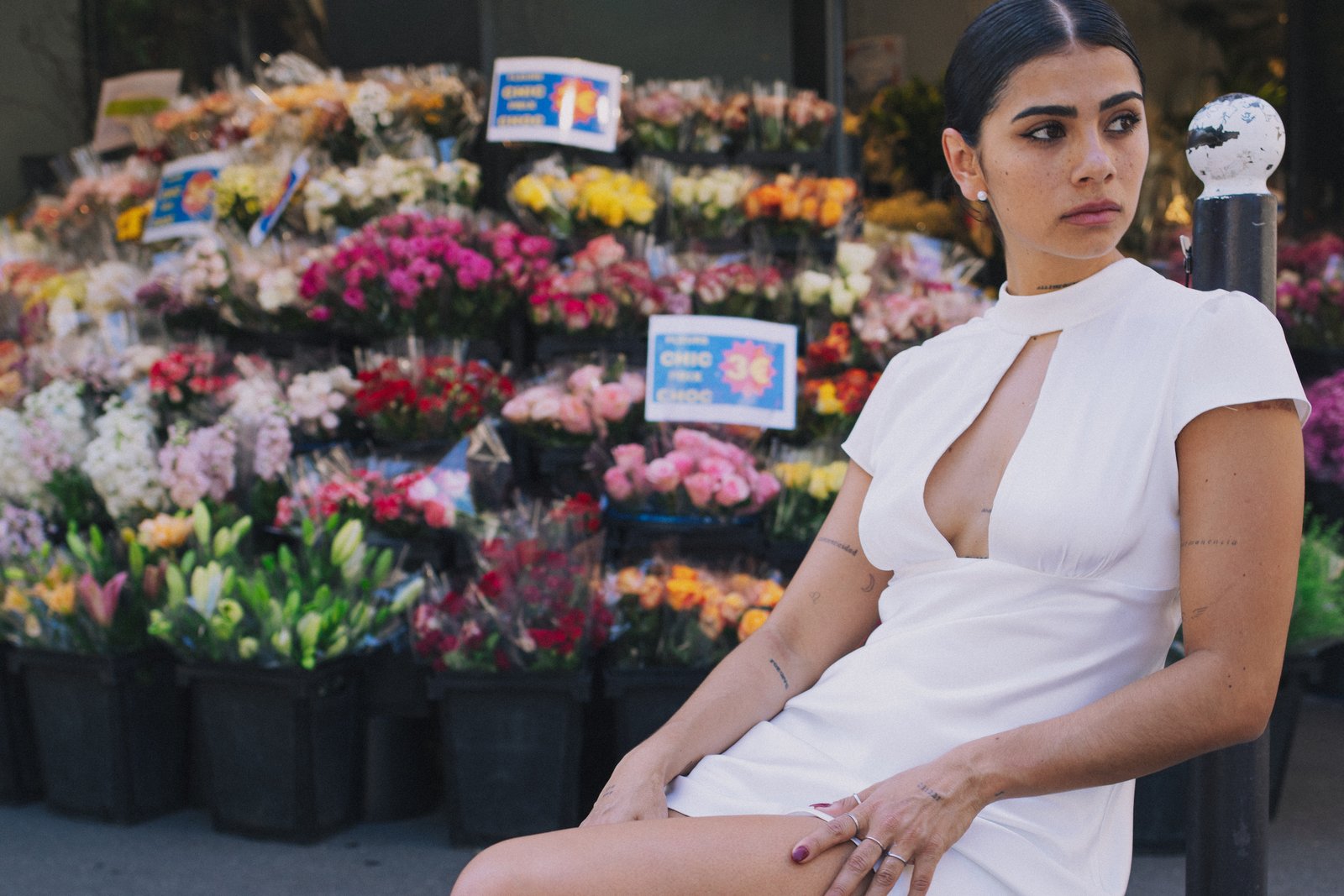 A woman in a white dress sits beside a florist's banked rows of flowers, looking off-camera.