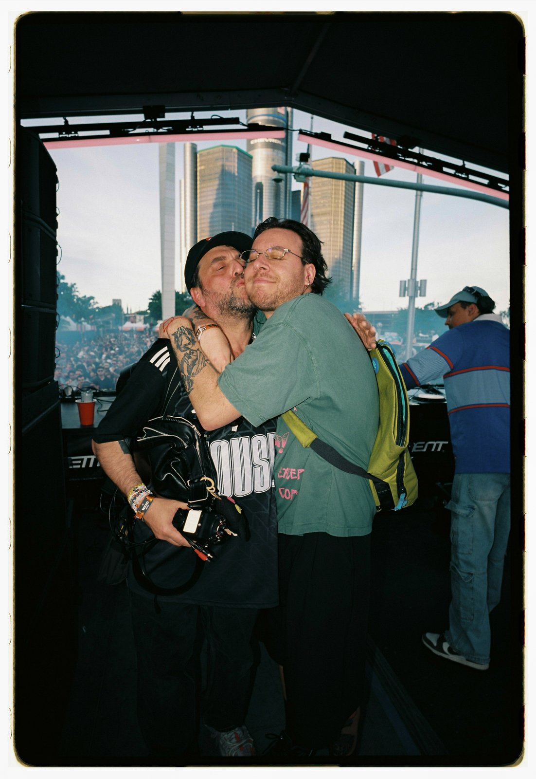 Two men embrace on a festival stage, city skyline and crowd visible behind, framed film print.