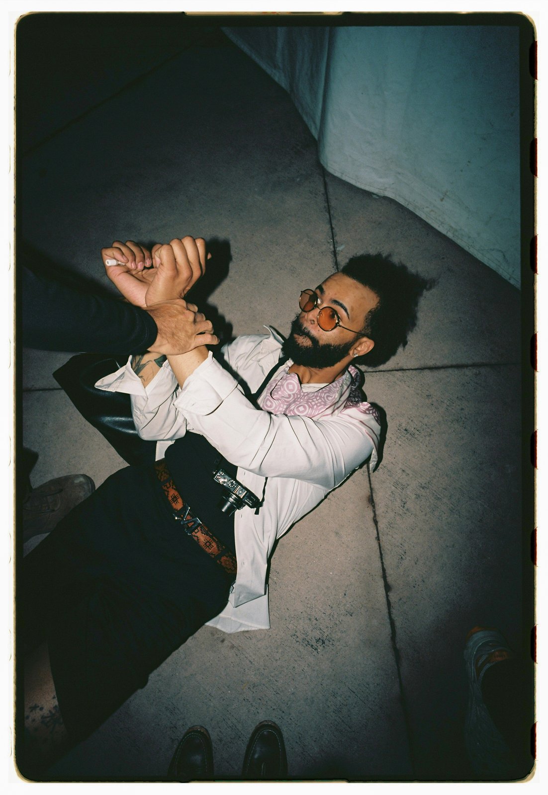 A bearded man in round sunglasses and white shirt lounges dramatically on concrete steps, flash-lit film with dark border.