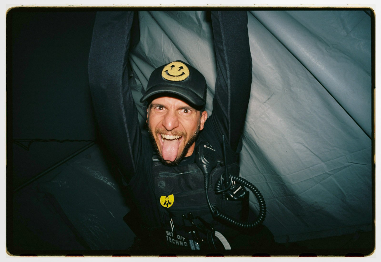 A man in a smiley-face cap and tactical vest grins with his tongue out inside a dark tent, flash film with white border.