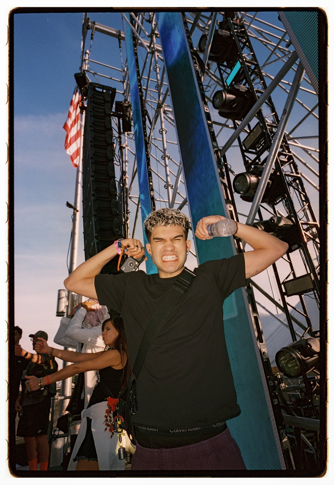 A young man in a black tee flexes both arms before festival stage rigging, an American flag and blue sky behind.