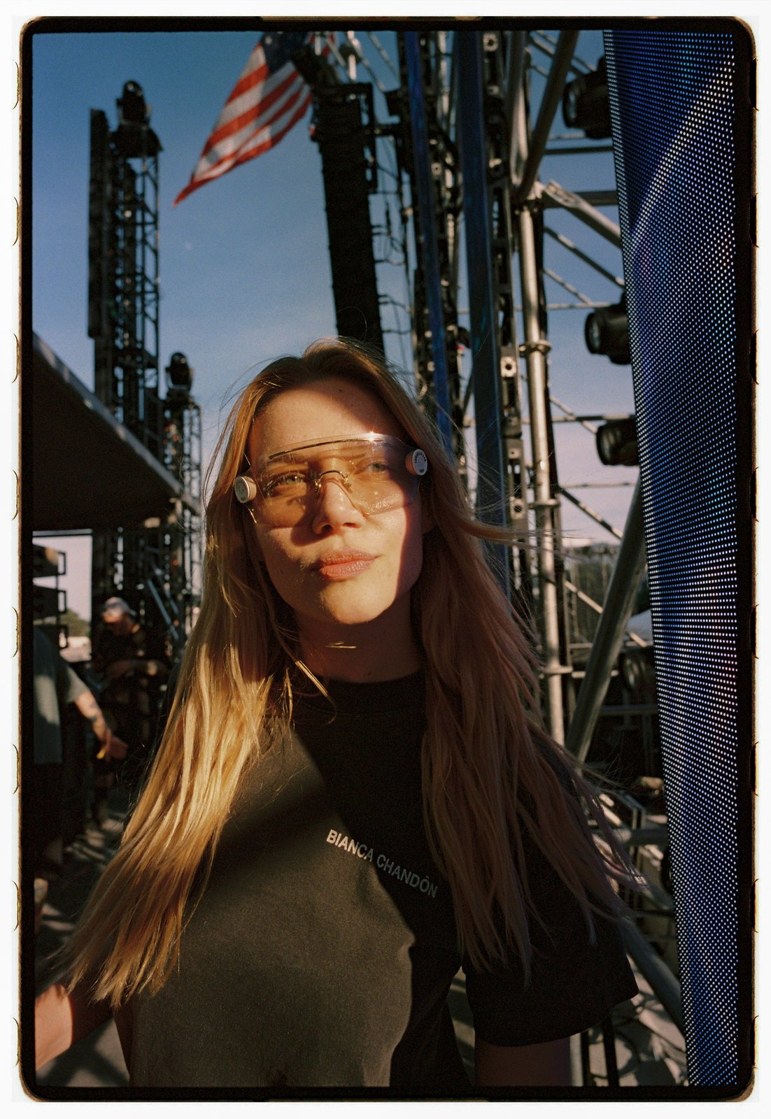 A long-haired woman in tinted glasses and a dark tee stands amid festival stage scaffolding in warm sunset light.