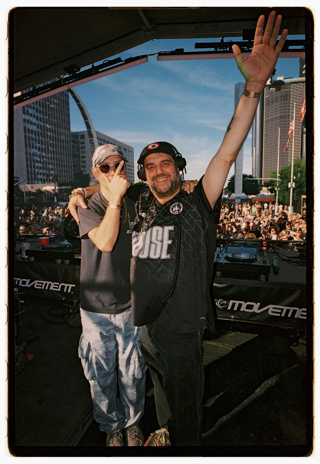 Two men pose behind DJ decks, one waving to a large outdoor crowd with a city skyline behind.