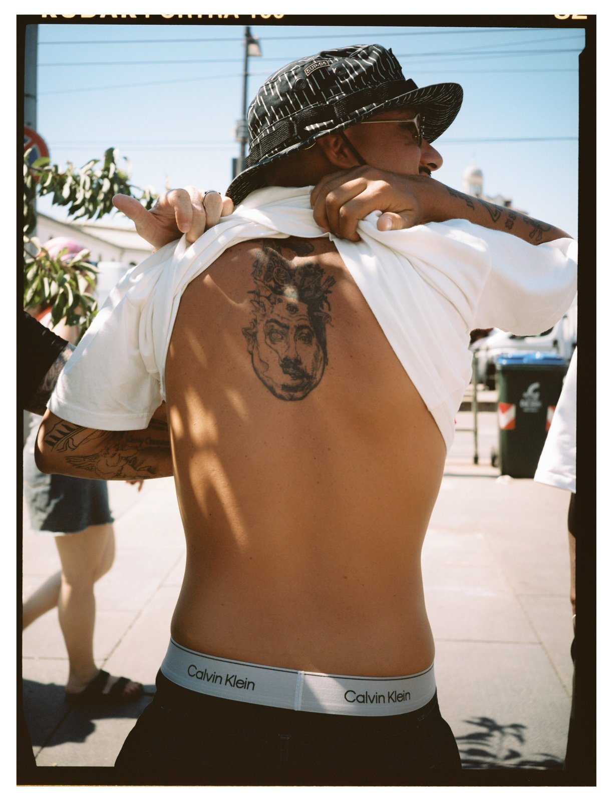 A shirtless man in a bucket hat shows a large back tattoo of a face on a sunny street.