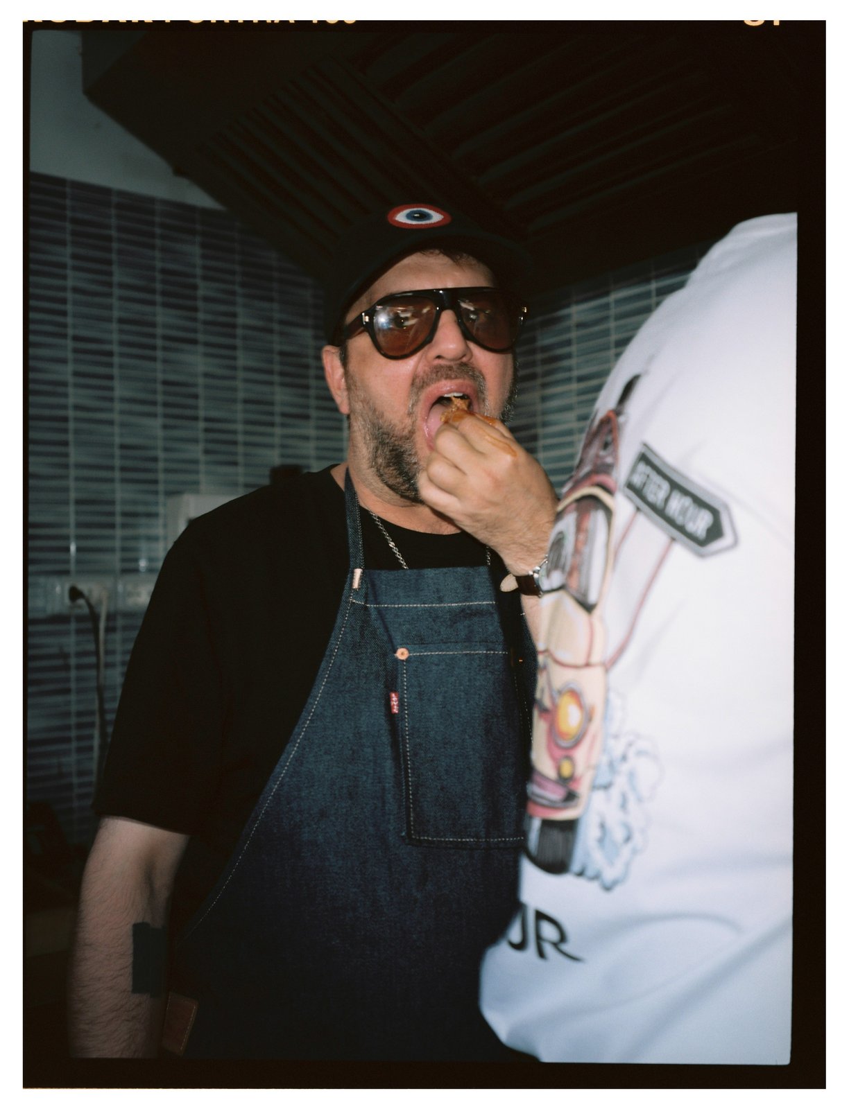 A man in sunglasses and a denim apron tastes food handed to him in a dim kitchen.