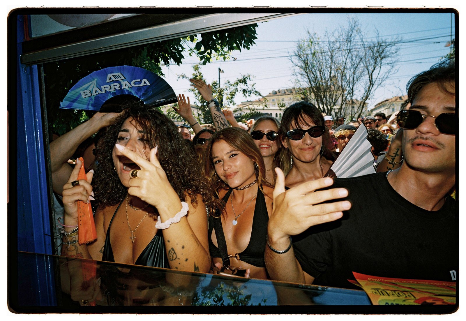 A crowd in sunglasses waves printed paper fans at a sunny outdoor event.