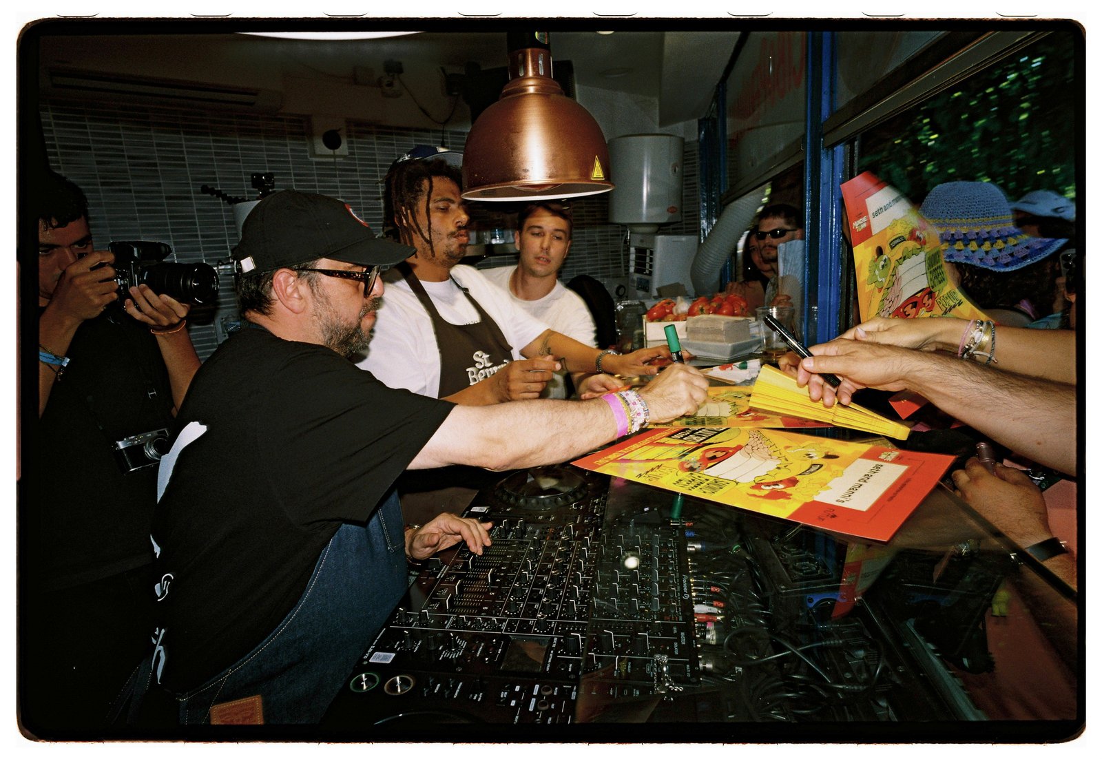 People reach across a counter to hand yellow flyers to a man at a DJ mixer while another films.