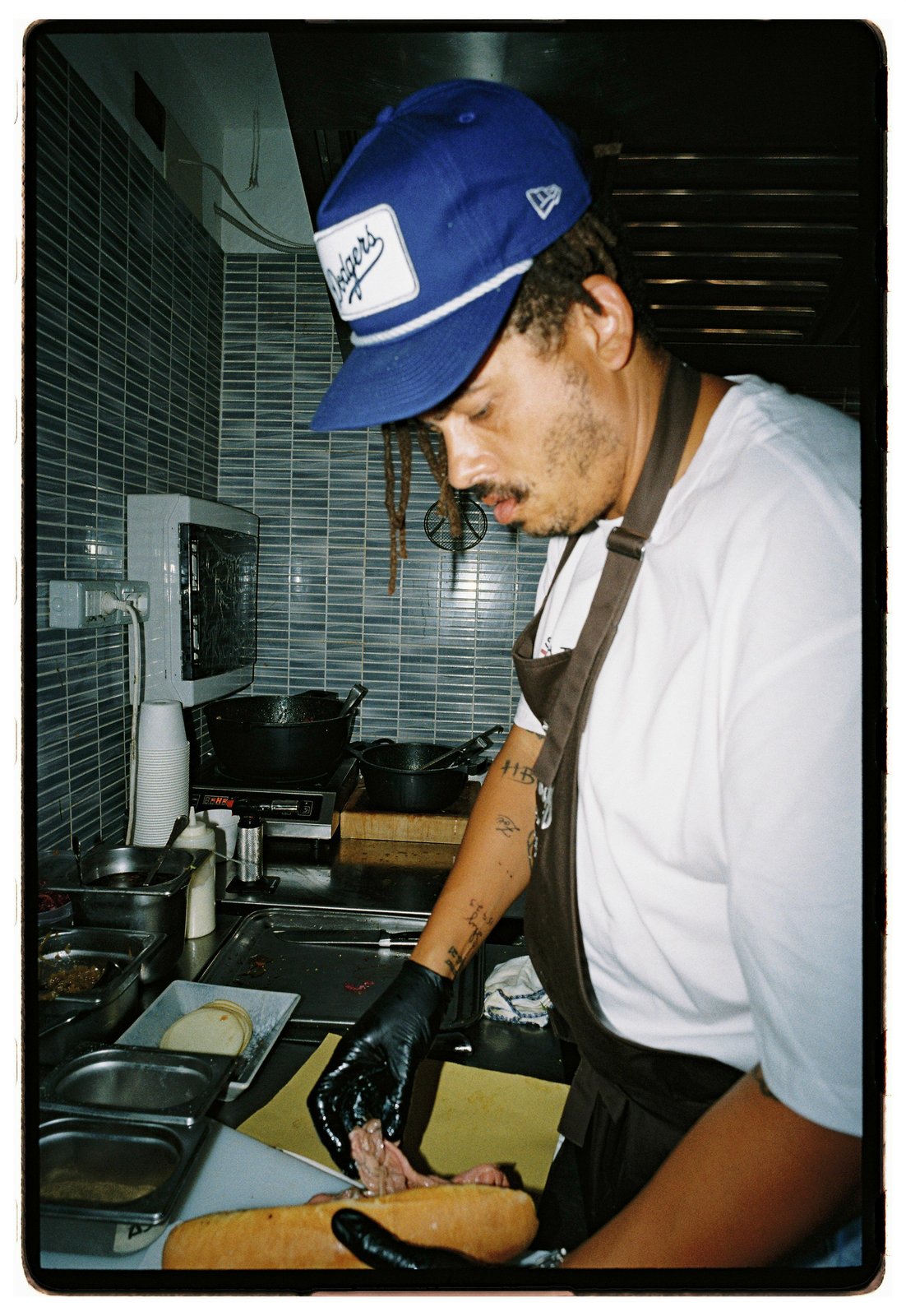 A man in a blue cap and apron with a gloved hand prepares food in a tiled kitchen.