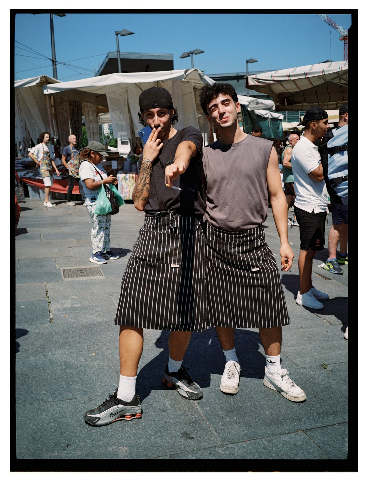 Two men in pinstriped aprons and sneakers pose at an outdoor market, one gesturing at the camera.