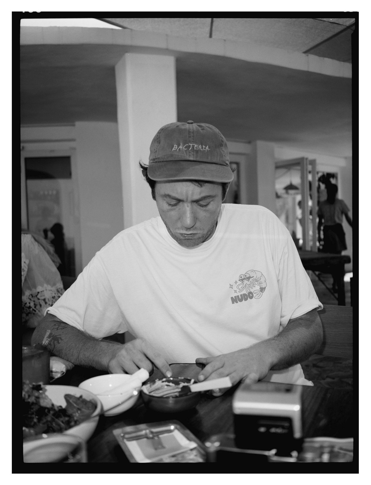 Black-and-white flash photo of a man in a cap and t-shirt eating at a table set with bowls.