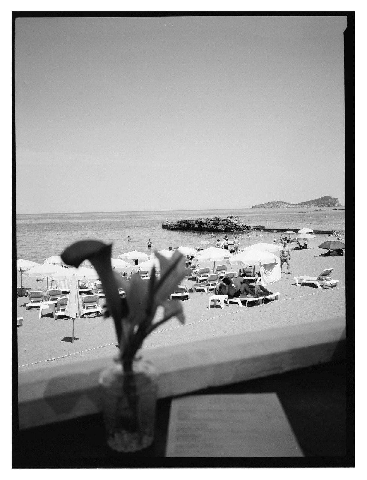 Black-and-white view past a flower vase to a beach of sun loungers, umbrellas and sea.