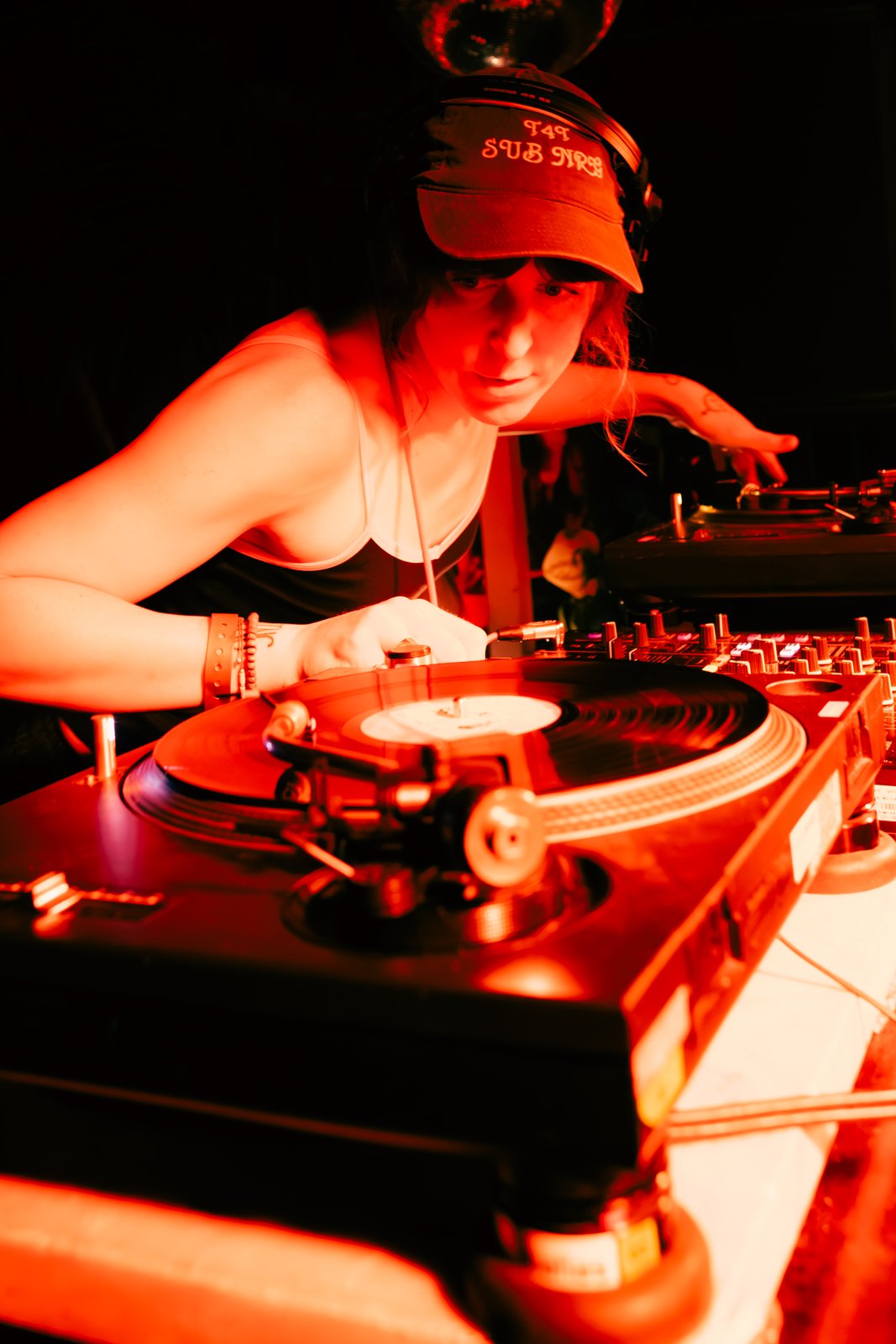 Bathed in red light, a DJ in a visor cap leans over vinyl turntables and a mixer in the dark.