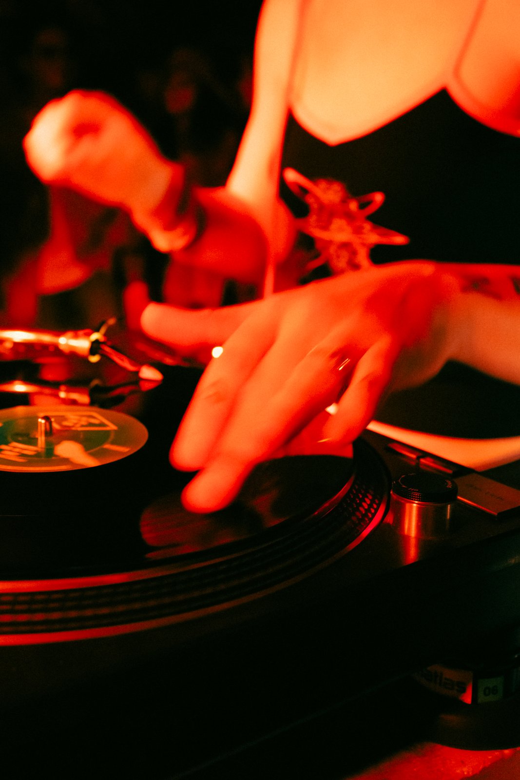 Under red light, a woman's hands work a spinning record on a turntable while a blurred figure moves behind.