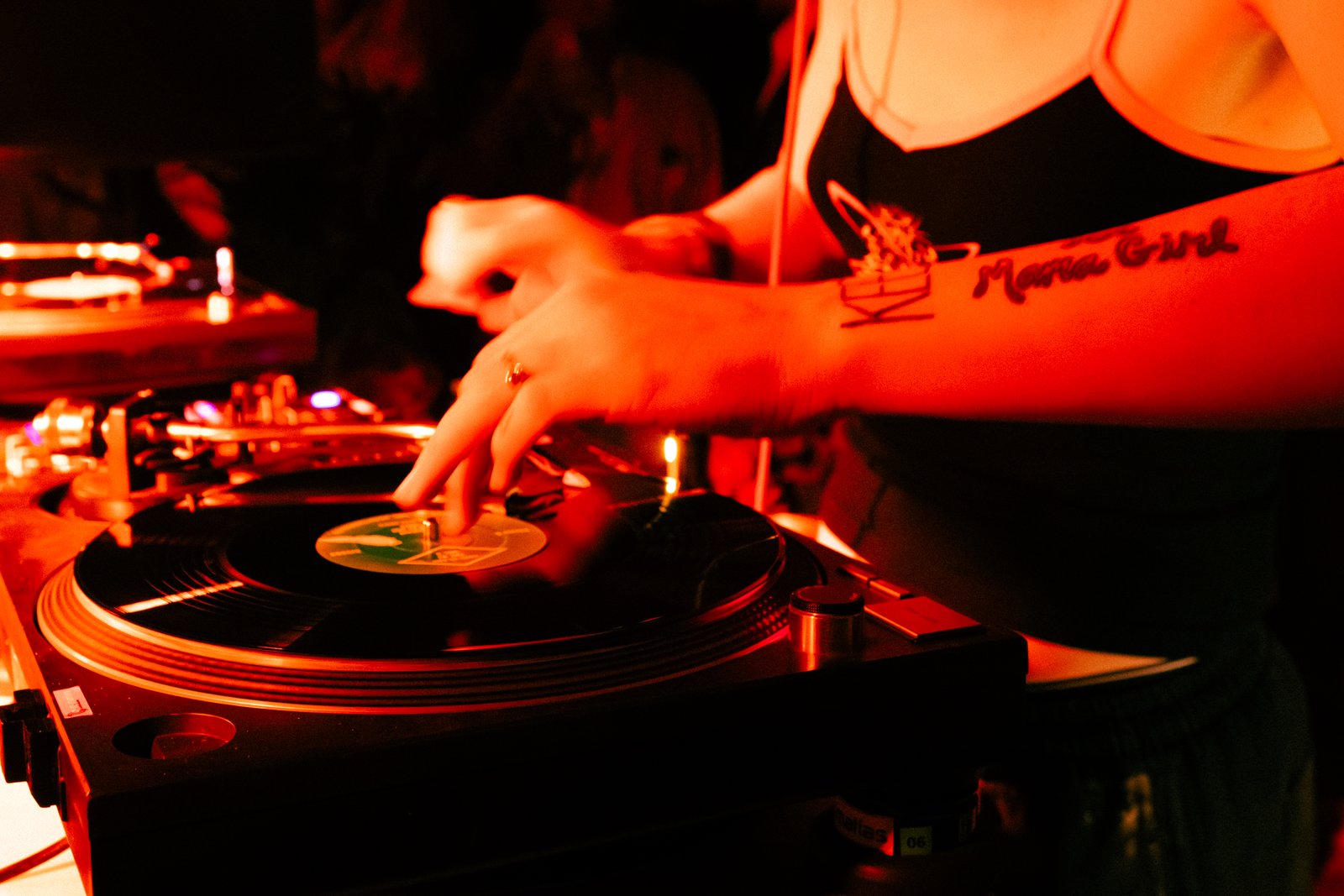 Close red-lit shot of a woman with a forearm tattoo cueing a record on a turntable, a second deck beside her.