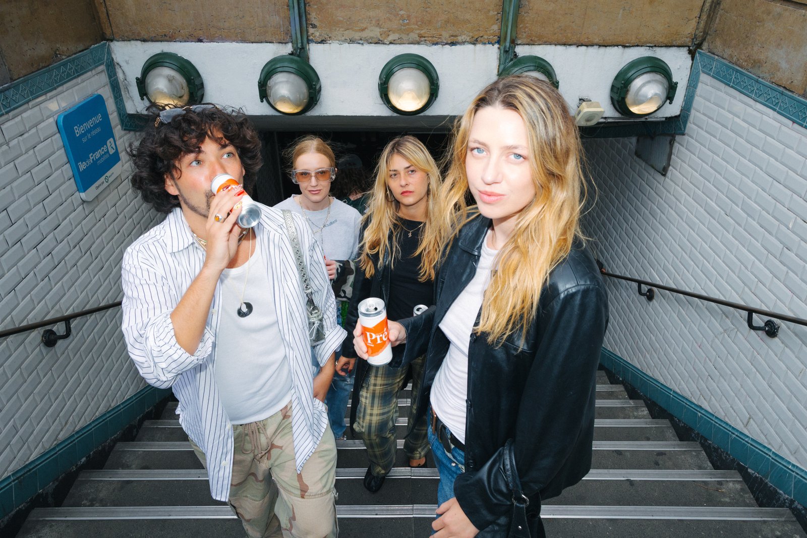 Four young people descend a tiled metro staircase toward the camera, one drinking from a can, under round station lights.