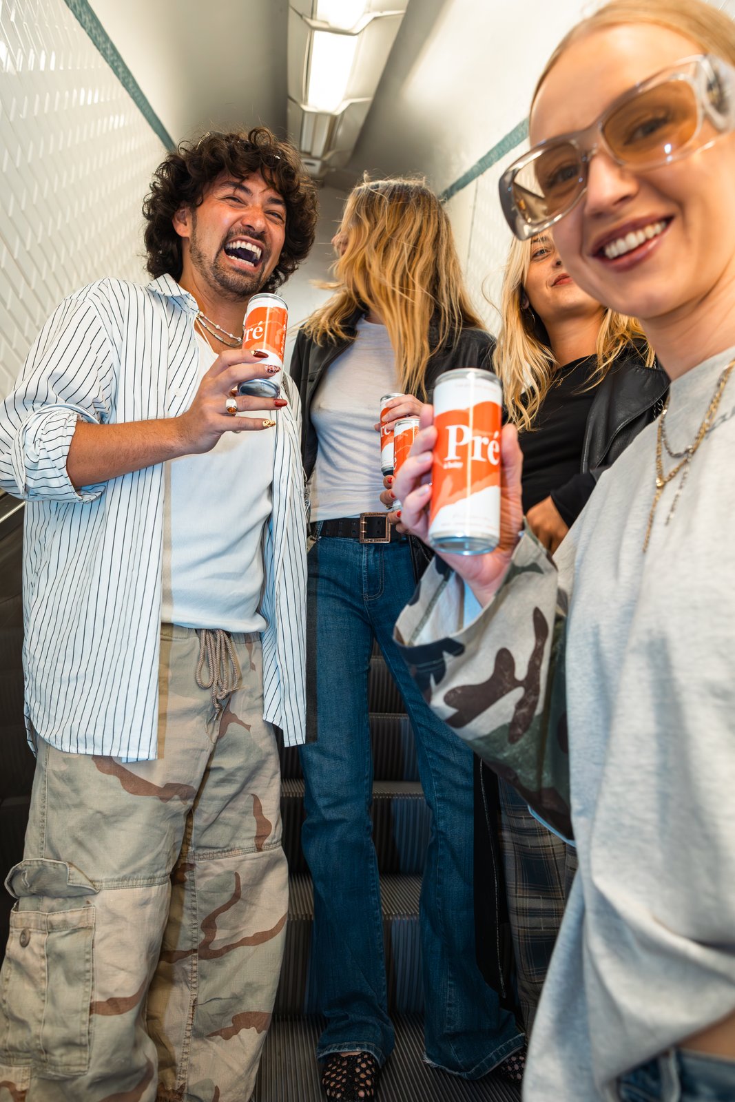 A laughing man and smiling woman hold orange cans on a metro escalator with friends behind them.