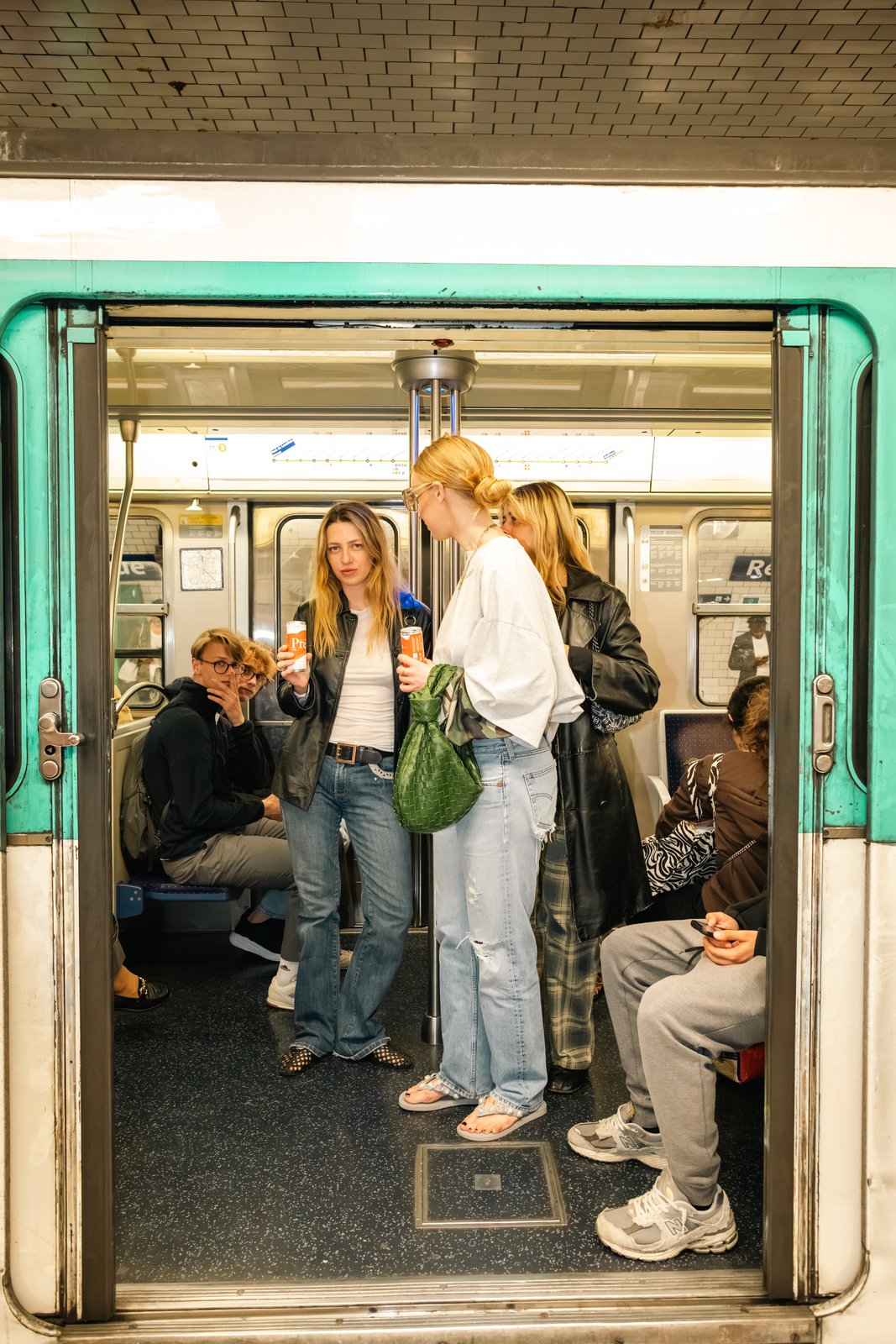 Two women holding drinks stand in the doorway of a green-and-white metro carriage with seated passengers inside.
