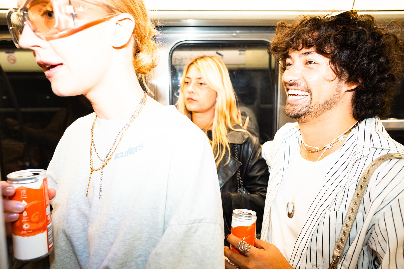 A smiling man and a woman in orange-tinted glasses hold orange cans aboard a metro train, warm flash light.