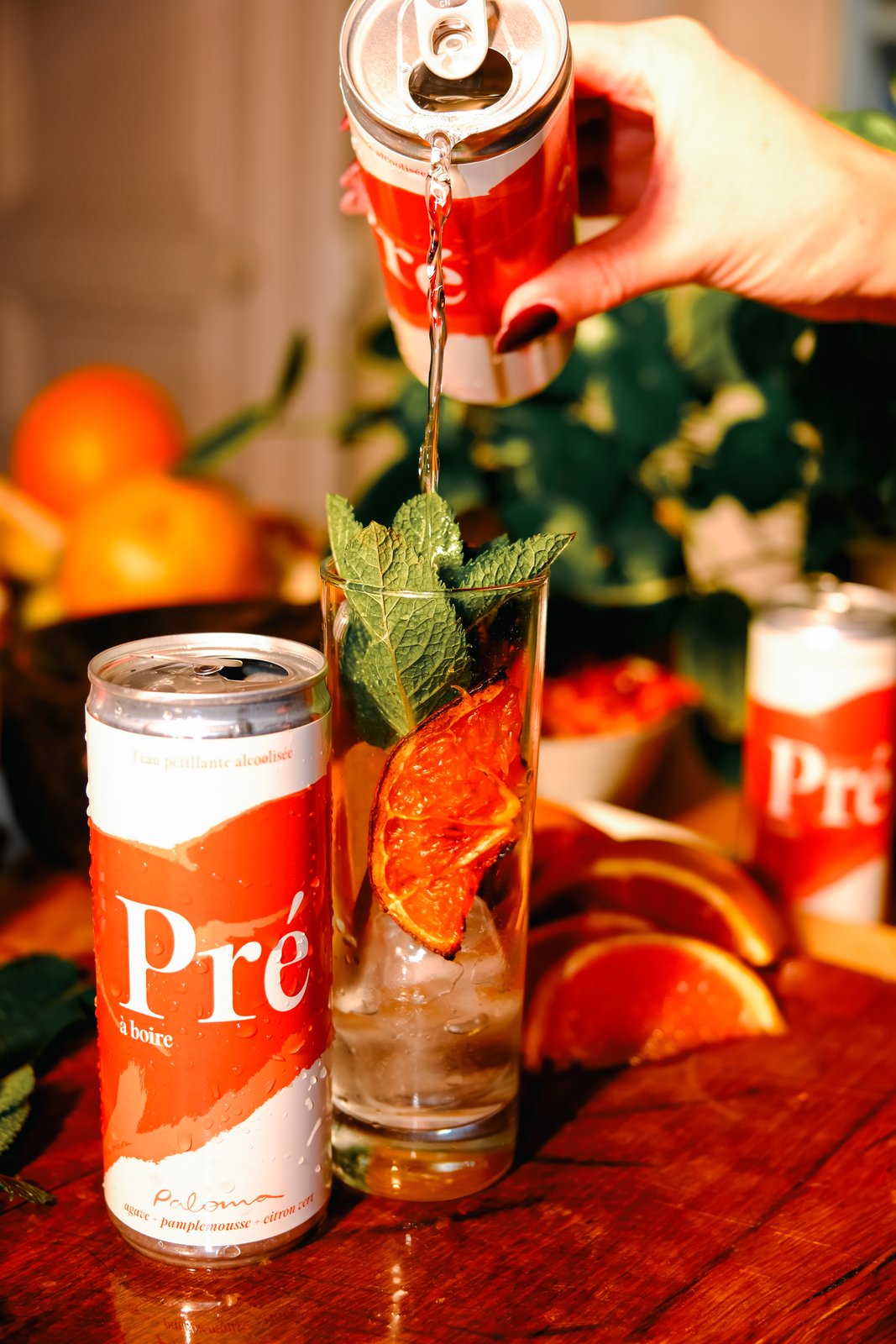 A hand pours from a red can into a tall glass with mint and dried orange, cans and citrus on a wooden table.