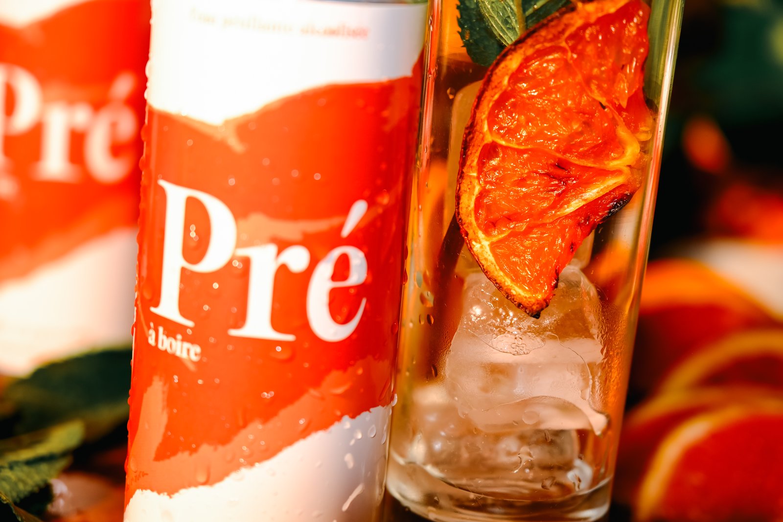 Close-up of a red-and-white drinks can beside a glass with ice and a dried orange slice, citrus in the background.