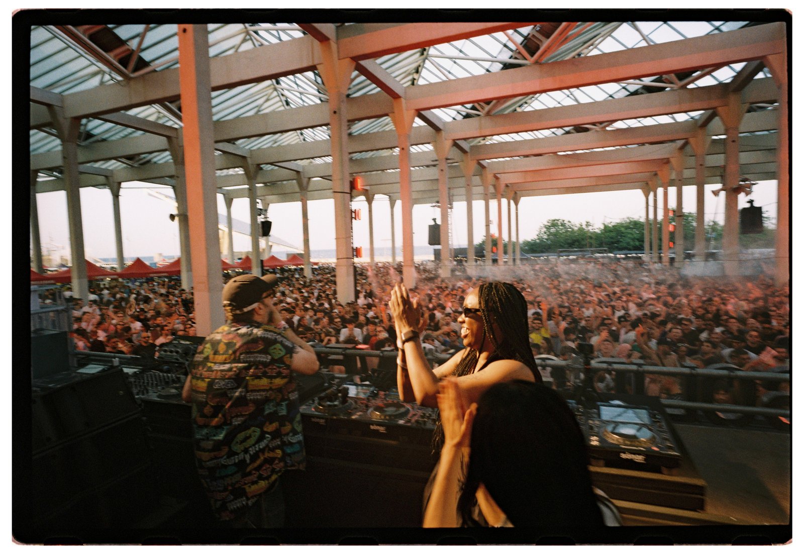 Two people clap onstage facing a huge sunlit crowd under an industrial glass-roofed venue.