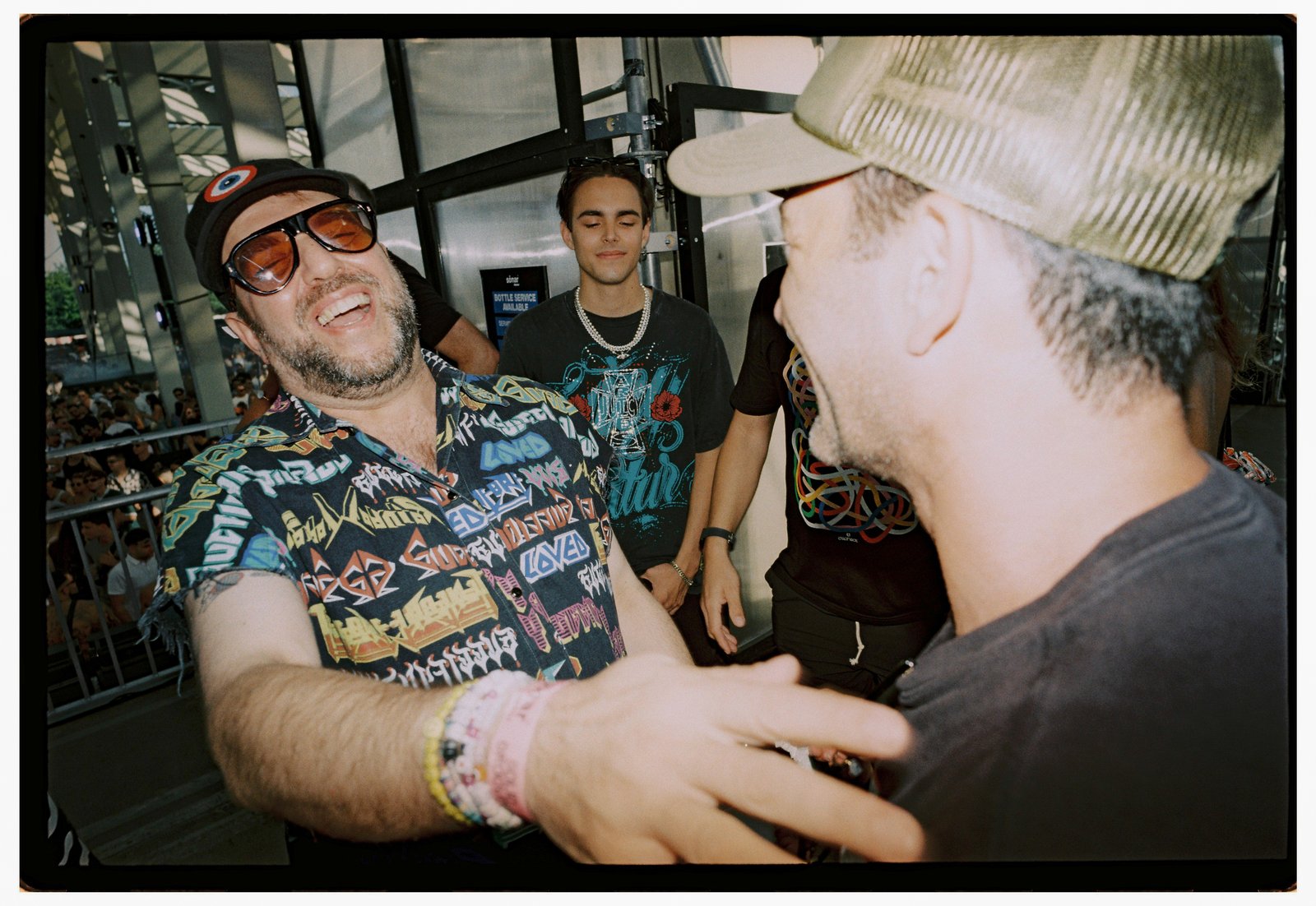 Laughing men greet each other outdoors, one in sunglasses and a patterned shirt, warm flash-lit film.