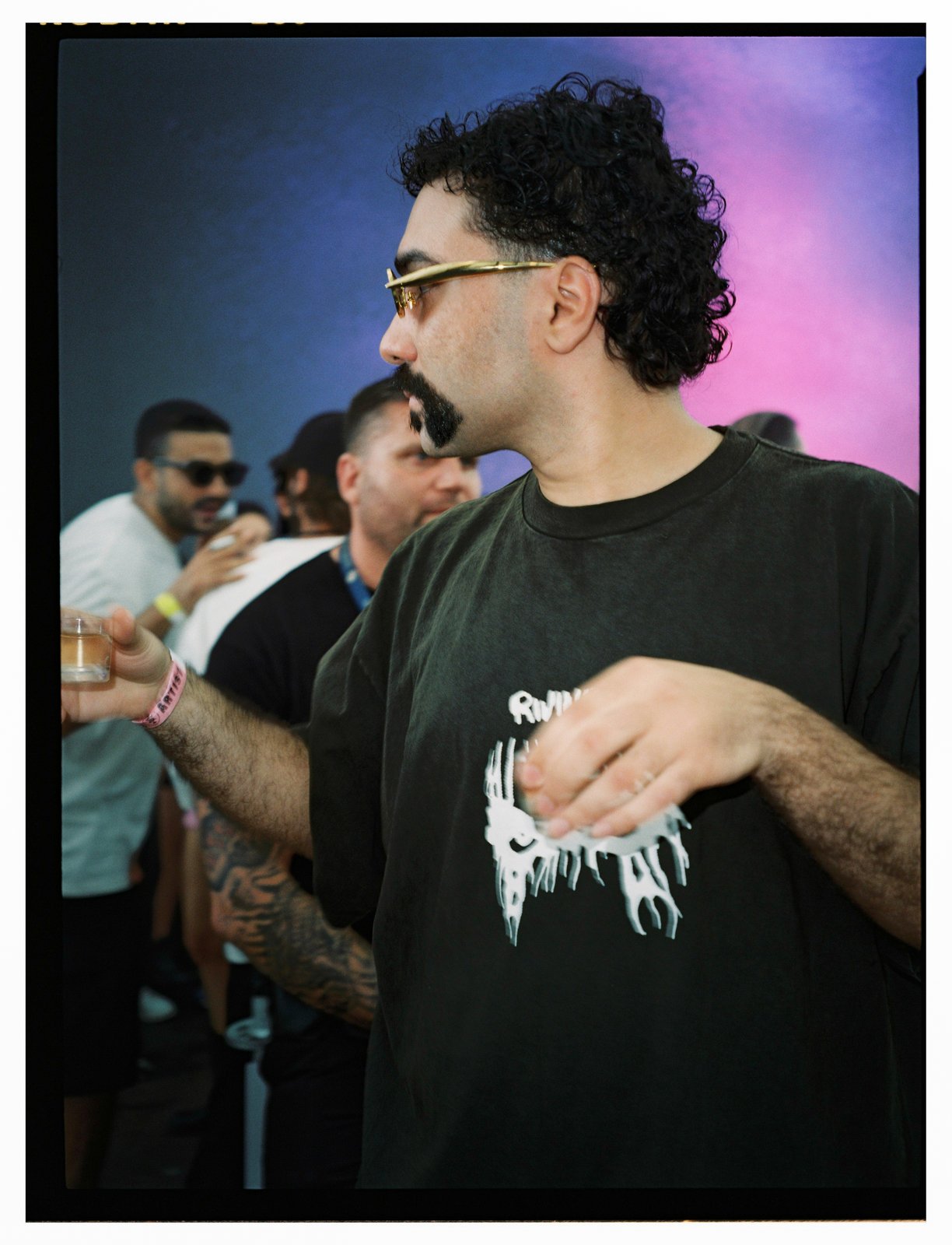 A curly-haired man in sunglasses and dark tee stands among partygoers against a pink-and-blue backdrop.