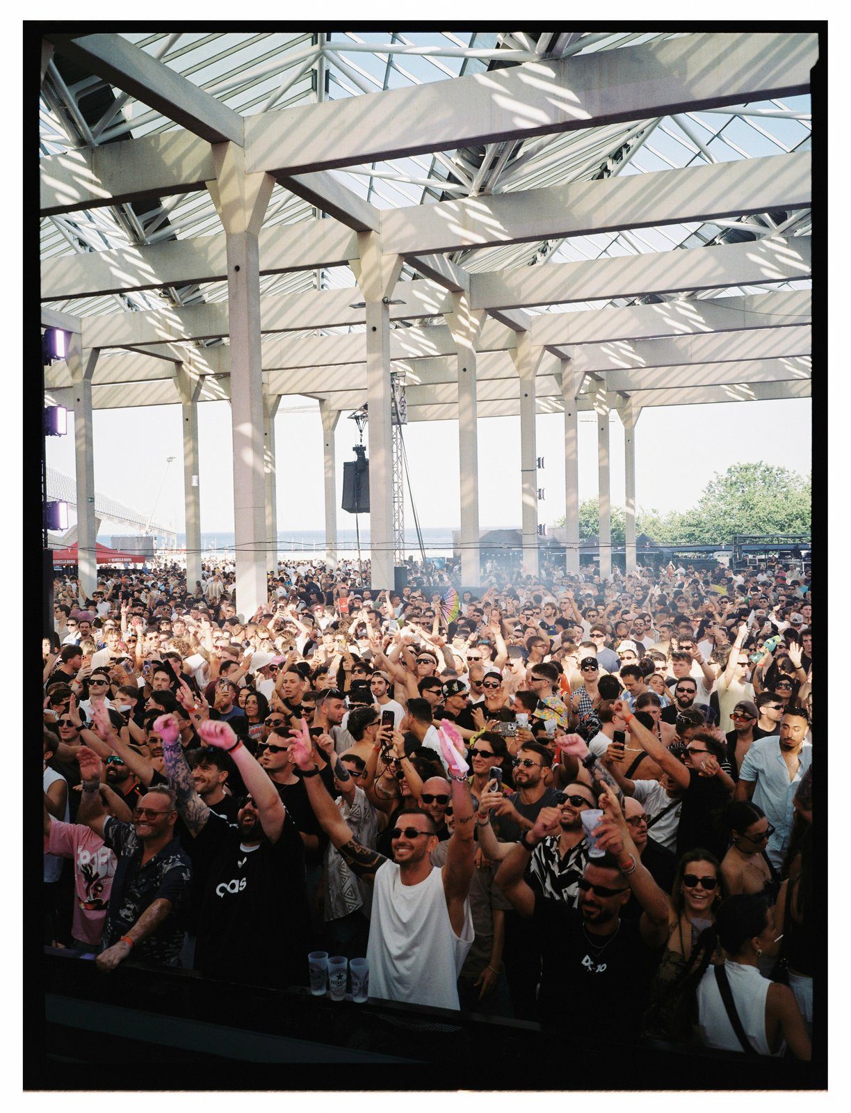 Dense daytime crowd with raised arms and phones under a bright glass-roofed open-air venue.