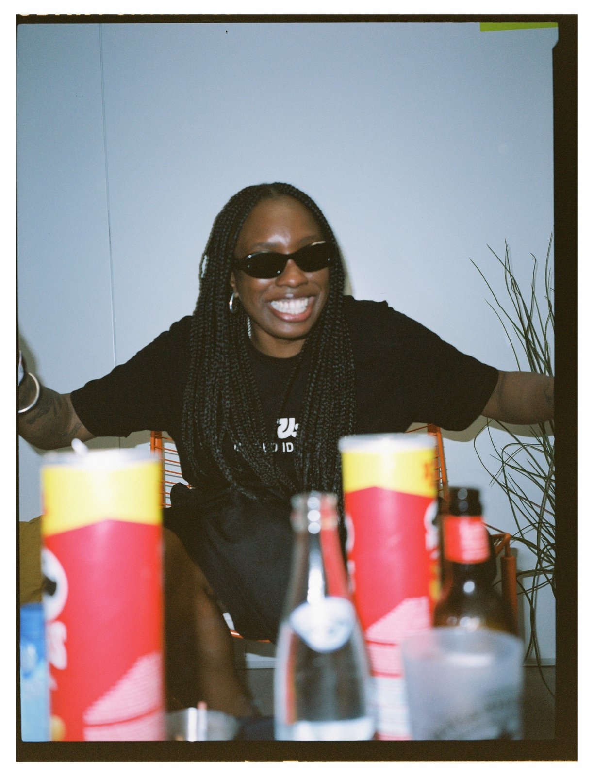 A grinning woman in sunglasses with long braids sits behind a table of drinks and cans.