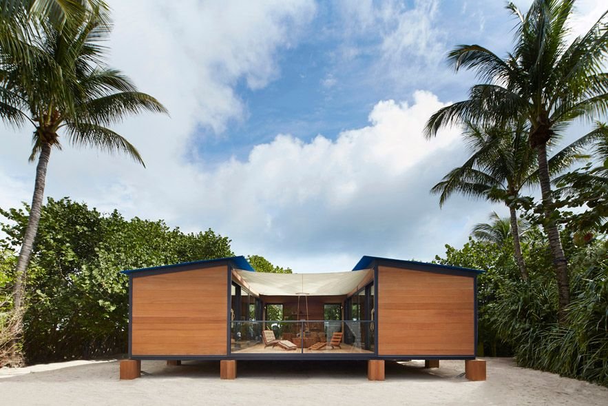 Wood and glass beach house with a tented roof set on sand between palm trees.