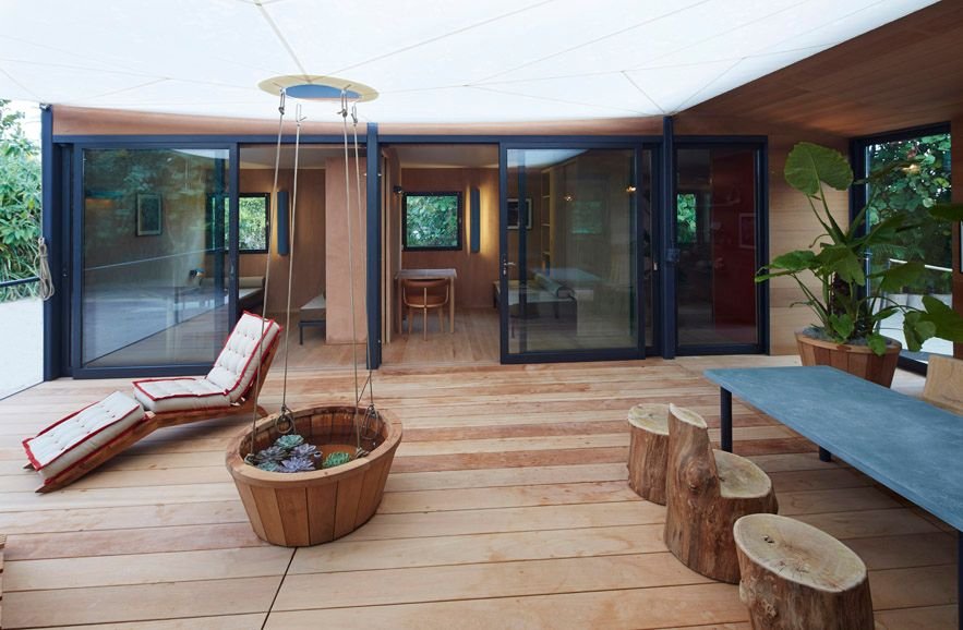 Wooden deck under a tented canopy with a lounge chair, log stools and a hanging planter.