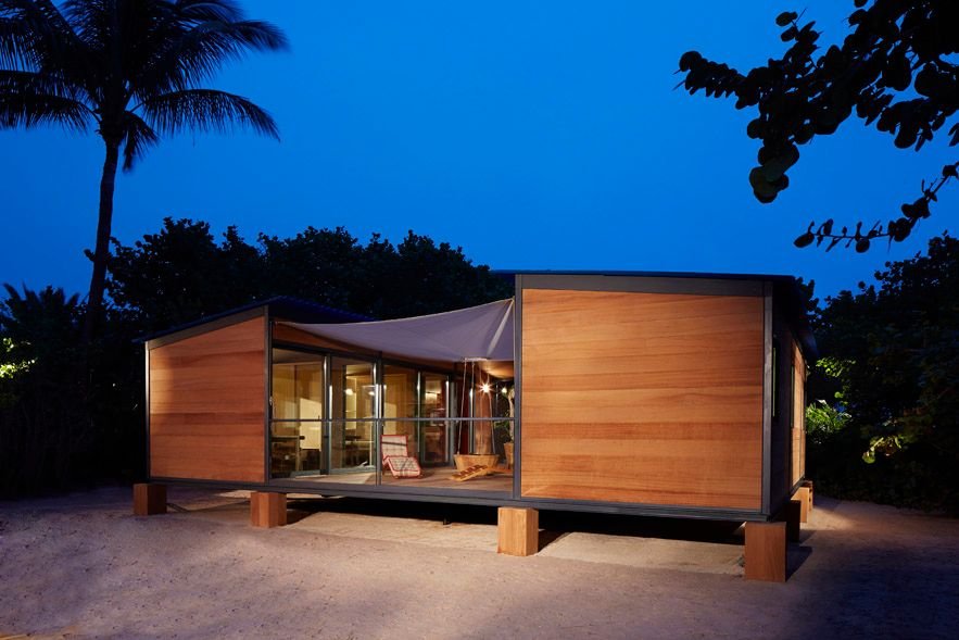 Illuminated wood and glass beach house on sand at dusk under a deep blue sky.
