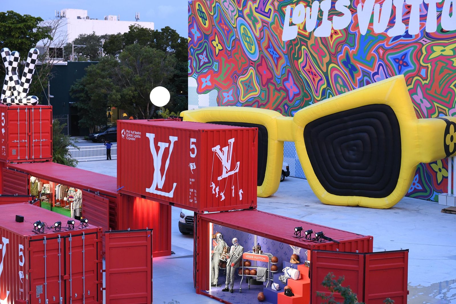 Outdoor display of stacked red shipping containers, giant yellow inflatable sunglasses and a psychedelic mural.