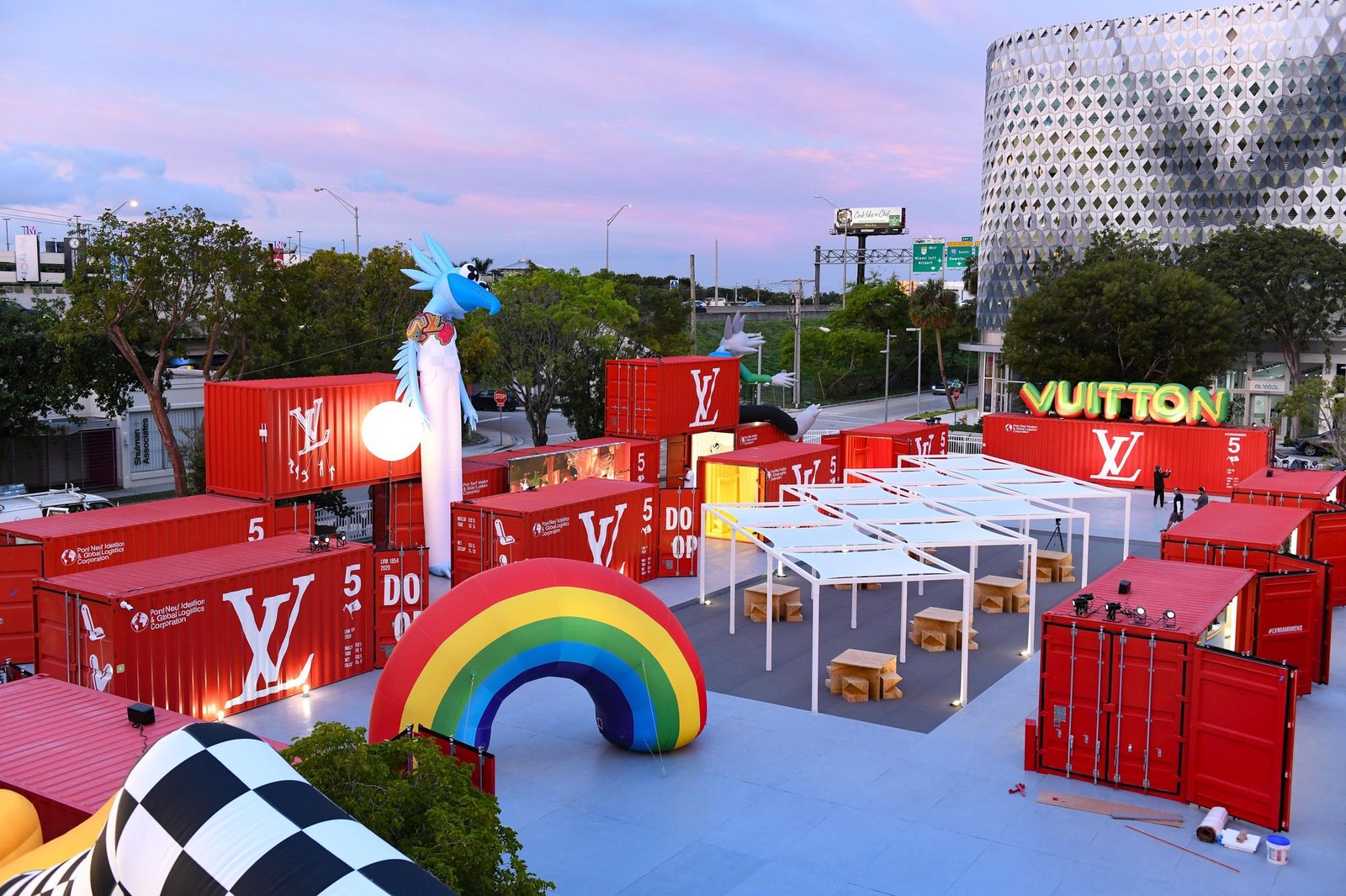 Dusk view of a red-container installation with a rainbow arch and glowing yellow "Vuitton" letters.