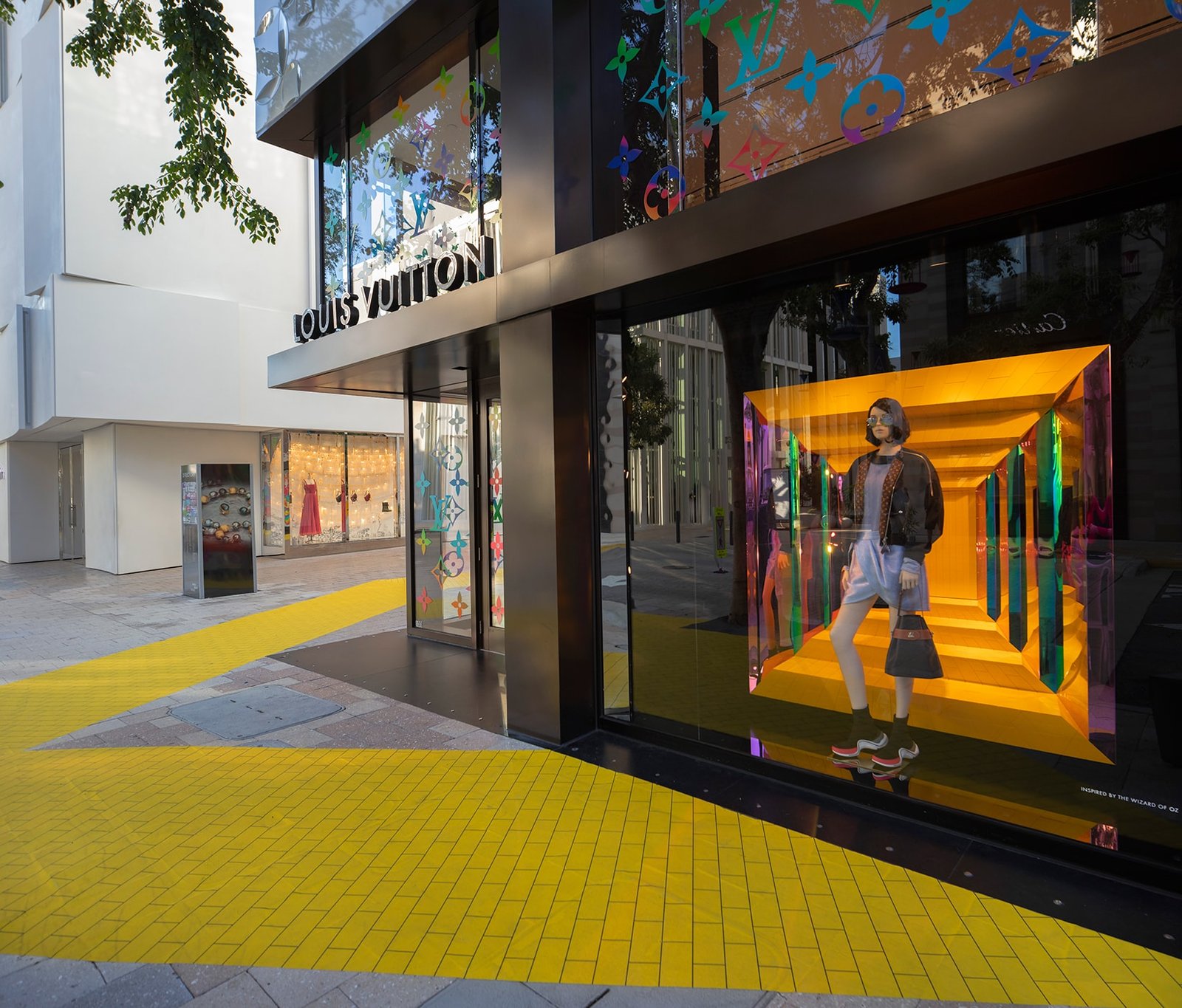 Window display with a mannequin inside a glowing orange mirrored box beside a yellow-tiled walkway.