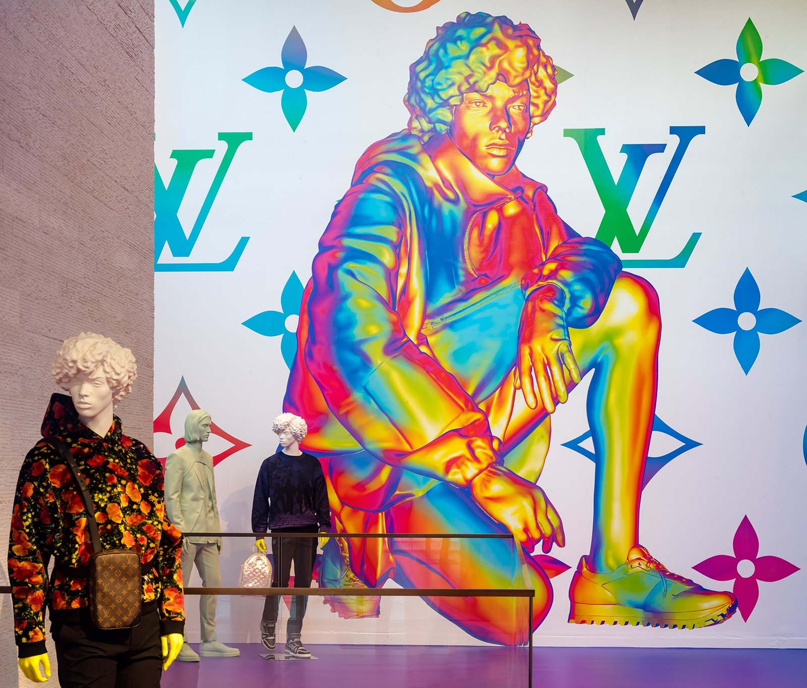 Mannequins in patterned menswear before a wall mural of a chrome rainbow crouching figure.