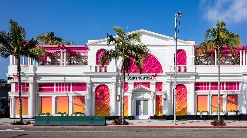 White two-storey building with arched windows in pink-to-orange gradients and palm trees along the street.